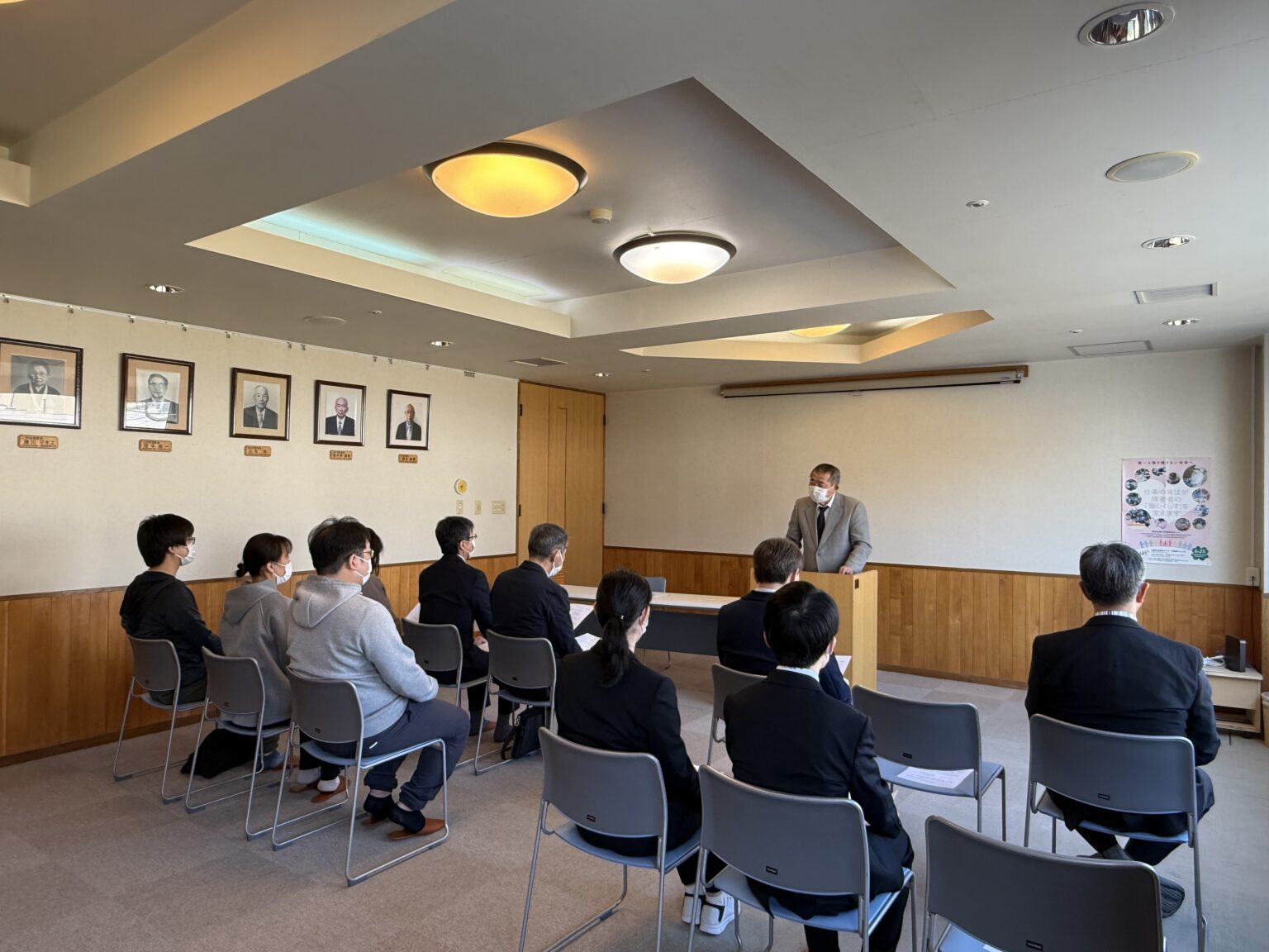 介護施設の共用部で行われるレクリエーションや講話の様子。壁に掲示された人物写真と照明が特徴。